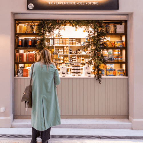 Woman in front of an artisan gastronomy shop – A woman standing in front of a specialty food shop selling artisanal Greek culinary products.