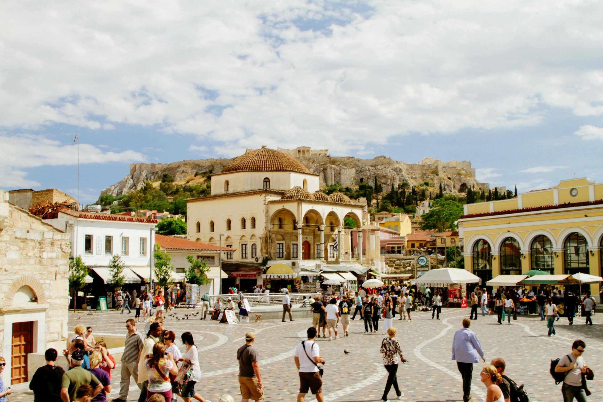 Monastiraki Square in Athens, Greece, featuring a vibrant market scene, the historic Tzistarakis Mosque, and the Acropolis in the background on a sunny day.